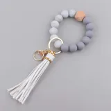 Llavero con pulsera de cuentas de silicona en tonos gris y blanco, con una cuenta de madera natural y borla blanca de flecos. Incluye aro metálico dorado y mosquetón para colgar llaves.