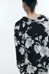 Camisa de sarga estampada con flores en blanco y negro, de corte recto, con cuello clásico, cierre frontal con botones, mangas largas con puños abotonados y bajo redondeado.