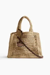 Bolso de mano tipo shopper, confeccionado en paja trenzada color beige, con detalles en cuero marrón. Incluye correa fina extraíble y etiqueta decorativa en un asa.