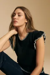Top tejido sin mangas color negro, con cuello redondo y volados en las sisas con terminación en contraste de punto blanco. Presenta un calce recto y puño inferior acanalado con detalle de punto blanco.
