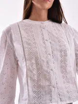 Camisa blanca con bordado calado, cuello redondo, mangas largas abullonadas con puños elásticos y cierre frontal con botones.