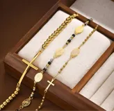Set de tres pulseras doradas con dijes religiosos: una de cuentas redondas con cruz grande, otra con cuentas negras y dijes de medallas, y otra con cuentas pequeñas y cruz pequeña.