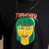 Remera negra de manga corta con estampado de una cara de mujer con pelo verde y llamas en los ojos, con el logo de Thrasher arriba.