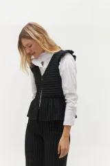 Chaleco blanco con finas rayas verticales negras, escote en V, cierre frontal con botones negros y volados en lino negro en los hombros. La parte inferior presenta otro volado en la misma tela del cuerpo.