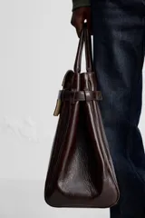 Bolso de mano extragrande color marrón oscuro, fabricado en piel de alta calidad. Tiene un compartimento principal con cierre magnético, un bolsillo interior con cremallera y un bolsillo secundario con solapa y cinta. Las asas son de piel.