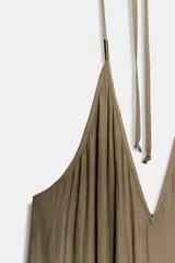 Mono largo de tejido satinado color verde oliva, con escote profundo en V y tirantes finos ajustables tipo halter que se anudan en la parte superior. Presenta una pieza metálica decorativa en el centro del escote y un bajo asimétrico con caída fluida.