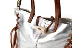 Mini cartera de cuero plateado con detalles en cuero marrón. Presenta flecos laterales largos, manijas superiores y un cierre frontal tipo cuerno. Incluye una correa larga ajustable y extraíble.