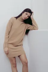 Vestido corto color beige de tejido de punto, con cuello alto tipo polera y mangas largas. Corte recto y terminaciones acanaladas en puños, cuello y ruedo.