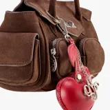 Bolso tipo satchel de gamuza color marrón, con dos bolsillos frontales con cierre y un bolsillo lateral con solapa. Cierre principal con broche metálico y dos asas de hombro delgadas. Incluye un charm colgante de cuero rojo con forma de cereza y un dije metálico dorado.