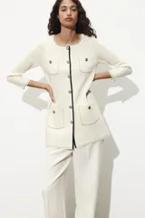 Chaqueta color crema de punto con cuello redondo y manga larga. Tiene bolsillos delanteros de plastrón, pespuntes combinados a contraste y cierre frontal con botones dorados.