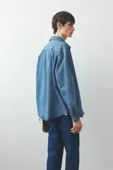 Camisa de denim azul de algodón rígido, corte holgado, cuello inglés, tapeta clásica con botones blancos, bolsillo de parche en el pecho, canesú en la espalda, mangas largas con puños abotonados y bajo levemente redondeado.