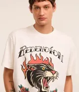 Remera color crema de corte recto, con cuello redondo y mangas cortas. Presenta un estampado estilo tatuaje con un felino en llamas y la frase "Redención es una virtud".