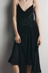 Vestido midi negro de tirantes finos con escote fluido y detalle de abalorios en el corpiño. Falda asimétrica con volados. Forro interior.