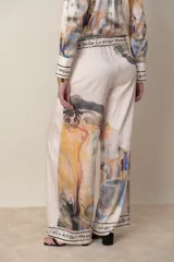 Pantalón palazzo italiano de satén de seda color natural con estampado en tonos de verde, lila y naranja. Cintura con elástico y cordón, bolsillos laterales. Cintura y bajo con guarda escrita.