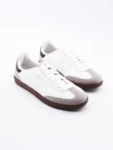 Zapatillas urbanas blancas con puntera de gamuza gris, suela marrón y detalle negro en el talón.