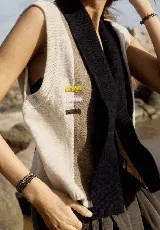 Chaleco de tejido de punto sin mangas, con diseño de bloques de color verticales en tonos beige claro, marrón topo y negro. Presenta un cuello chal negro y tres pequeños bordados horizontales en amarillo, lila y marrón en el frente.