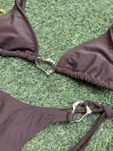 Conjunto de bikini color marrón chocolate, con corpiño triangular y bombacha colaless con tiras laterales para atar. Ambas piezas cuentan con argollas metálicas decorativas en el centro y los laterales.