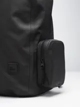 Mochila negra con cierre superior y bolsillo frontal con cierre. Tiene dos bolsillos laterales con cierre y un pequeño logo cuadrado en la parte inferior.