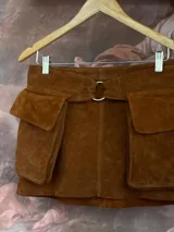 Falda corta de gamuza color marrón, con diseño utilitario que incluye grandes bolsillos cargo a los lados y un cinturón removible con hebilla metálica circular.