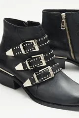 Bota corta de cuero negro con diseño inspirado en el estilo rocker. Presenta tres tiras frontales con remaches y hebillas metálicas color níquel, puntera metálica y taco ancho de 4 cm con detalle de vira metálica. Posee cierre lateral interno.