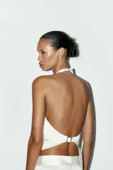 Top chaleco blanco de tejido satinado con cuello halter ajustable con hebilla metálica, falsos bolsillos de doble vivo en delantero, espalda descubierta con tira ajustable y cierre frontal con botones.