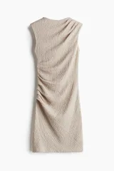 Vestido corto color beige, sin mangas, de punto con textura arrugada. Presenta cuello elevado y drapeado en un hombro y en un lateral de la cintura.