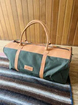 Bolso de lona verde con detalles en cuero color suela, cierre superior con cremallera y doble asa de mano.