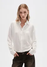 Camisa blanca de viscosa con cuello clásico, cierre frontal con botones y mangas largas. Presenta un sutil diseño de líneas verticales con strass. Corte al cuerpo.