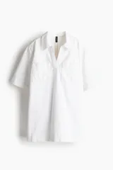 Vestido corto blanco de denim rígido, corte recto y holgado, con cuello camisero, escote en V, mangas cortas y bolsillos de parche en el pecho.