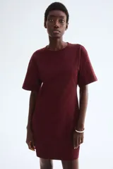 Vestido corto en punto de algodón suave color borgoña, con cuello redondo ribeteado acanalado, hombros caídos y mangas al codo. Pinzas moldeadoras en la parte posterior de la cintura para estilizar la figura.