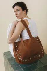 Bolso tote de cuero color marrón con apliques de tachas plateadas. Tiene doble asa de hombro y sobre interior de cuero.