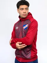 Campera de entrenamiento con capucha, cierre frontal y puños y cintura de rib. El cuerpo central es de tela estampada con patrón abstracto en tonos rojos y grises oscuros, mientras que las mangas y los laterales son de color rojo liso. Lleva el escudo del Club Nacional de Football y el logo de Umbro en el pecho.