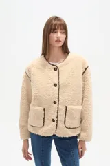 Campera de poliéster con textura de corderito en color beige, con cuello a la base y cierre frontal de botones. Presenta ribetes en contraste de color marrón oscuro en el cuello, la cartera, los hombros y en sus dos bolsillos frontales tipo parche.