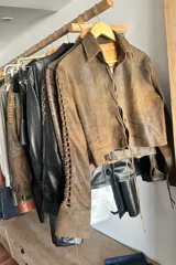 Camisa corta marrón de cuero ovino desgastado con cuello clásico, cierre frontal con botones y mangas largas con aberturas.