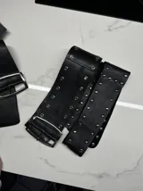 Cinto de cuero negro ancho de 9 cm, con ojalillos metálicos a lo largo del contorno.
