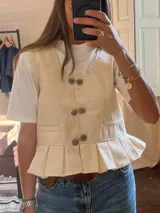 Chaleco color crema con cuello redondo, cierre frontal con botones metálicos y peplum con pliegues.