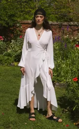 Vestido blanco de crepe con escote en V, mangas largas abullonadas con elástico en los puños, largo irregular, lazo para ajustar en la cintura y volado en el ruedo.