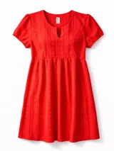 Vestido corto de broderie color rojo, con escote en V y pequeña abertura frontal, mangas cortas abullonadas y bolsillos laterales. Presenta un diseño de corte imperio con falda acampanada y textura calada característica del tejido broderie.