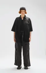 Camisa de manga corta color negro, confeccionada en popelín de algodón, con silueta oversize y dos bolsillos frontales, uno con solapa y otro con ojal y botón.