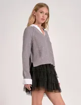 Buzo de tejido de punto gris, con cuello en V y detalles de lentejuelas. Se lleva superpuesto a una camisa blanca con cuello y puños visibles.