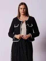 Chaqueta de tweed blanca con ribete negro, escote a la base, abertura central con botones, bolsillos delanteros y puños con aplique de botones perlados.