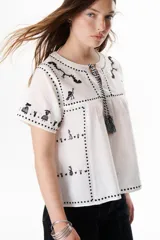 Blusa negra con bordados blancos de animales y motivos geométricos. Tiene cuello redondo con lazo y mangas cortas.