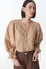 Camisa color beige con textura, escote en V, mangas largas abullonadas con lazos en los puños y cierre frontal con botones.