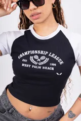 Remera corta blanca con mangas raglán negras, cuello redondo y estampado frontal con el texto "Championship League Est. 1996 West Palm Beach" y dos raquetas de tenis cruzadas.