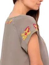 Blusa color taupe de mangas cortas con bordados florales en amarillo y fucsia. Tiene escote redondo con lazo y borlas amarillas.