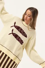 Sweater color manteca de tejido de punto con cuello redondo y ribete marrón. Presenta dos peces marrones tejidos en el frente con detalles bordados a mano.