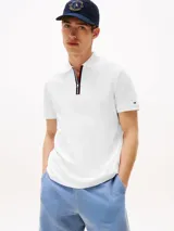 Chomba blanca de piqué de algodón, corte regular, con cuello tipo polo y cierre de cremallera con cinta distintiva en rojo, blanco y azul. Logo de Tommy Hilfiger bordado en la manga.