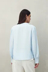 Camisa celeste de manga larga con cuello redondo y escote en V. Presenta detalles de pasamanería en el frente y las mangas, y botones forrados al tono. De corte fluido.