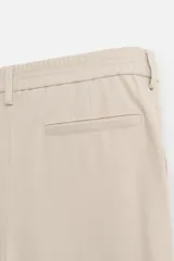 Pantalón gris de corte ancho con cintura elástica, pliegues frontales, bolsillos laterales y traseros de vivo. Incluye detalle de hebilla metálica en la trabilla y cierre frontal con cremallera y botón.