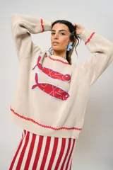 Sweater tejido color crudo con cuello redondo y mangas largas. Presenta dos peces rojos con detalles en azul bordados en el frente. Tiene ribete ondulado rojo en cuello, puños y ruedo.